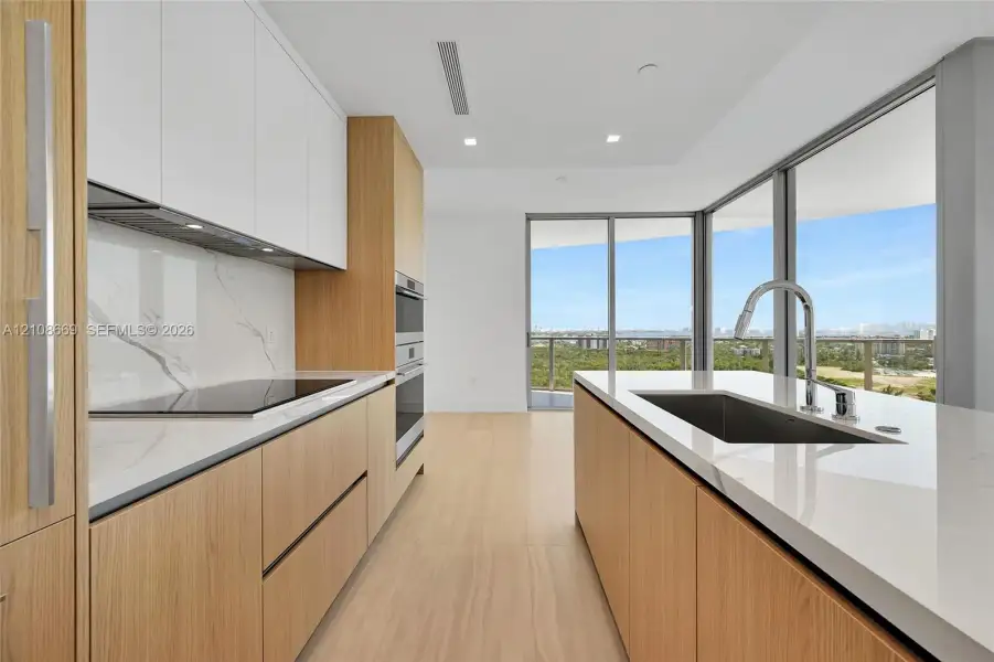 Modern kitchen featuring wood-finish cabinetry, white stone countertops, a full-height stone backsplash, stainless steel built-in double ovens, and floor-to-ceiling glass windows