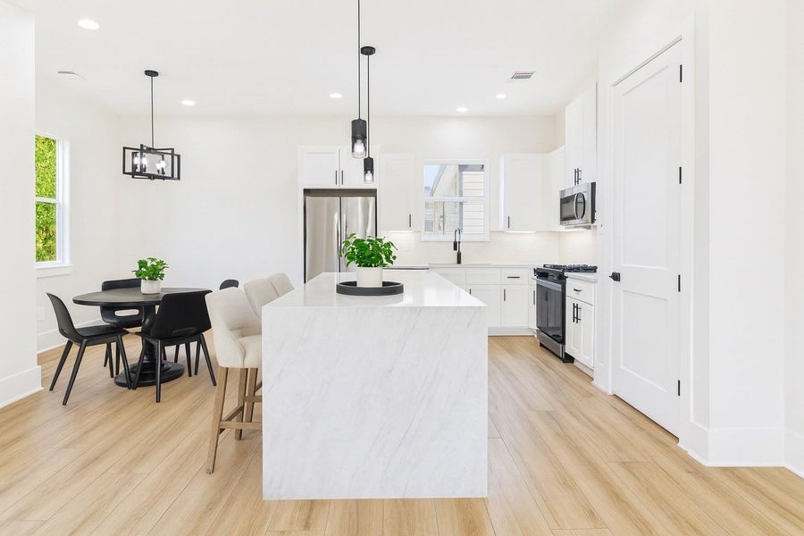 This modern kitchen features sleek white cabinetry, a spacious marble island with seating, and stainless steel appliances. It has an open layout with a dining area, light wood flooring, and ample natural light, creating a bright, welcoming atmosphere.