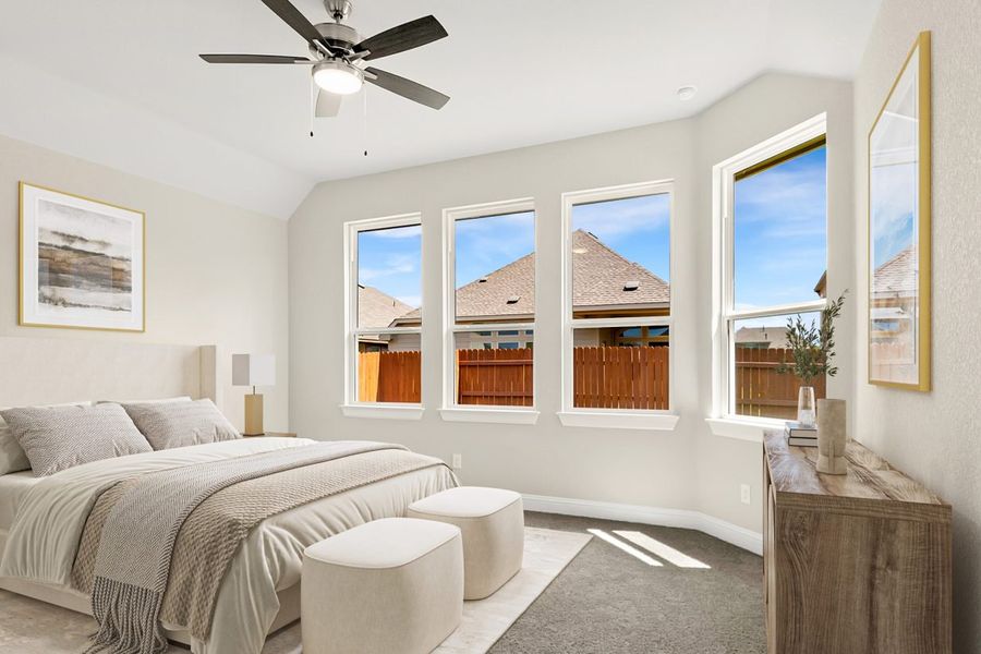 Image of a staged interior primary bedroom with a beige queen sized bed with beige bedding and wall decor. The walls are a painted a cream color with grey carpeting and four large windows
