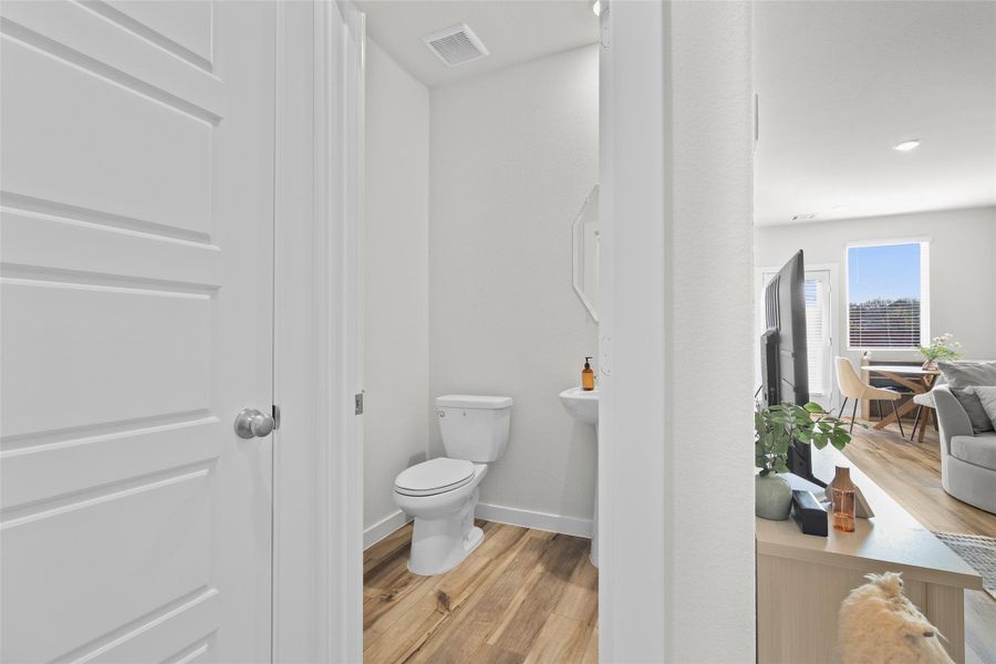 This photo features a compact bathroom with a toilet and pedestal sink. It opens into a bright living area with wood flooring, a cozy seating arrangement, and a dining space near large windows.