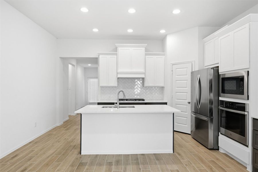 Sleek modern kitchen with bright white cabinetry, stainless steel appliances, and a central island featuring a built-in sink, all under recessed lighting and set against a chic herringbone tile backsplash.
