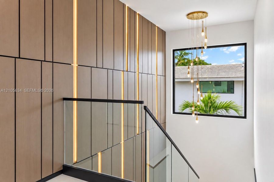 Custom wood-panelled accent wall with integrated vertical lighting, glass railing system featuring a black metal handrail, and multi-pendant brass chandelier