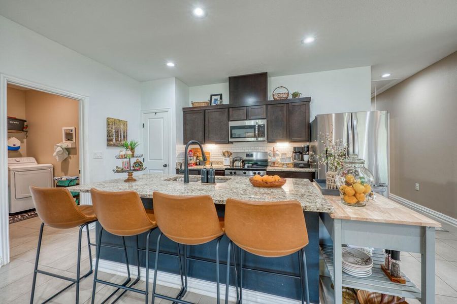 Spacious kitchen featuring an expansive granite-finish island with seating, dark wood-finish cabinetry, stainless steel appliances, and a subway tile backsplash