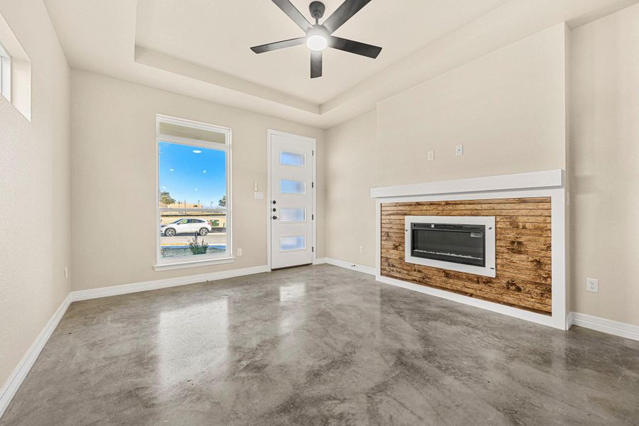 Unfurnished living room with finished concrete flooring, a ceiling fan, a glass covered fireplace, and a tray ceiling