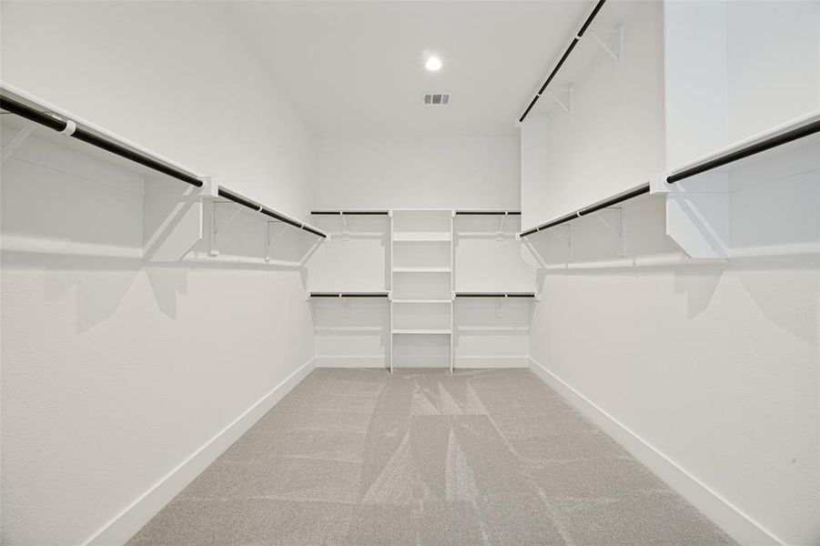 Spacious walk-in closet featuring white built-in shelving, black hanging rods, and light gray carpet flooring Spacious walk-in closet featuring white built-in shelving, black hanging rods, and light gray carpet flooring