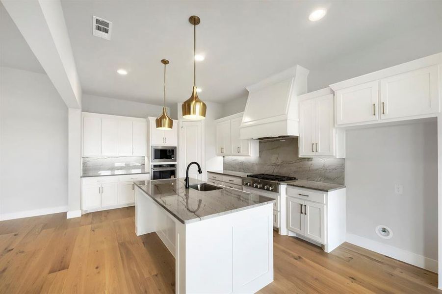 Gourmet kitchen featuring a central island with a sink, brass pendant lighting, white cabinetry, a built-in oven and microwave, and a gas range with a decorative hood