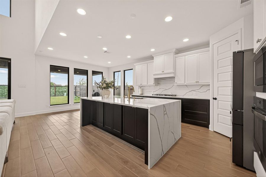 This modern kitchen features a spacious layout with sleek white cabinetry, a large marble island, and stainless steel appliances. The open design is enhanced by ample natural light from large windows, complemented by warm wood flooring.