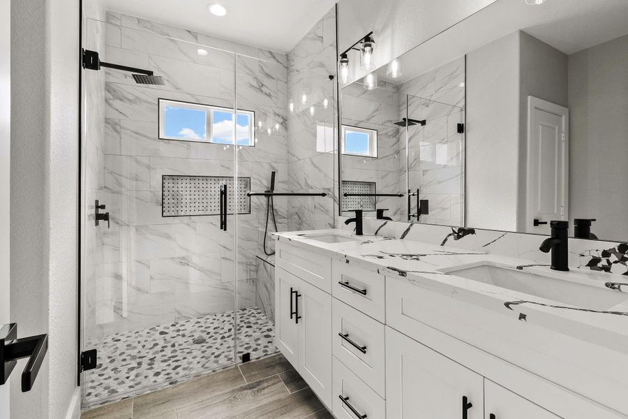 This modern bathroom features a spacious glass-enclosed shower with marble-like tiles and a dual vanity with sleek black fixtures. The design is bright and elegant, with ample lighting and contemporary finishes.