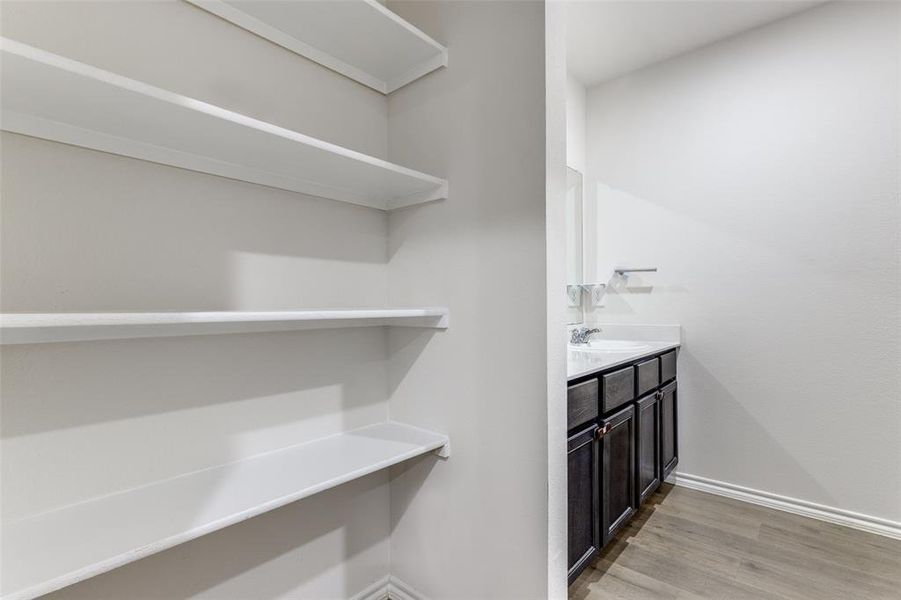 White shelving system with three levels, wood-finish flooring, a dark wood-finish vanity with cabinet storage, a white countertop, and a wall-mounted mirror White shelving system with three levels, wood-finish flooring, a dark wood-finish vanity with cabinet storage, a white countertop, and a wall-mounted mirror