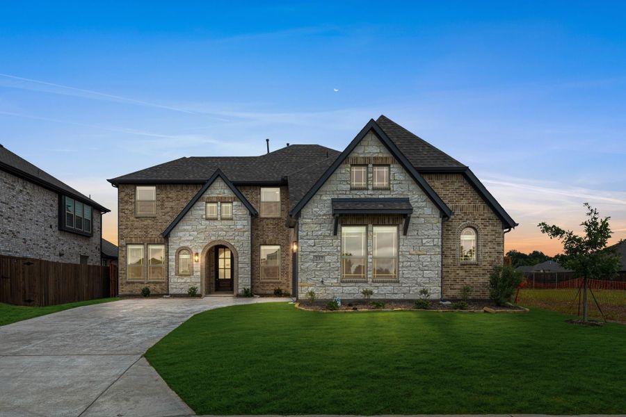 Two-story brick and stone home with arched entry, dark roof, and curved driveway at dusk