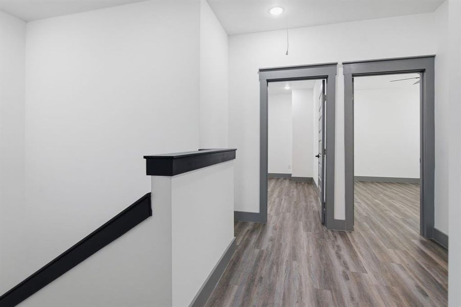 Hallway featuring wood-finish flooring, two panel doors with dark trim, recessed lighting, and a partial staircase with a dark railing