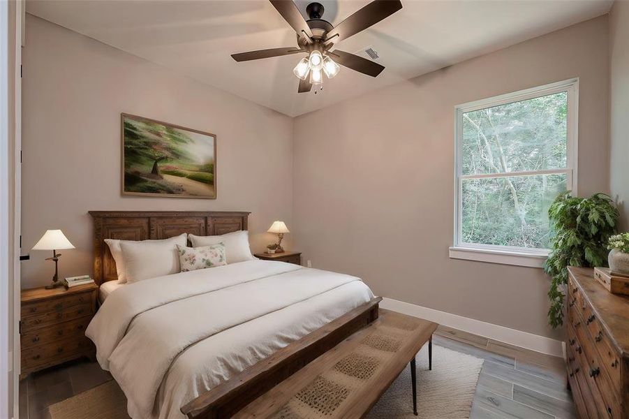 This is a cozy bedroom featuring a queen-sized bed with a wooden headboard, matching bedside tables with lamps, and a bench at the foot of the bed. The room is decorated with a large landscape painting above the bed and has a serene view of greenery from the window. It's finished with a ceiling fan and light fixture.