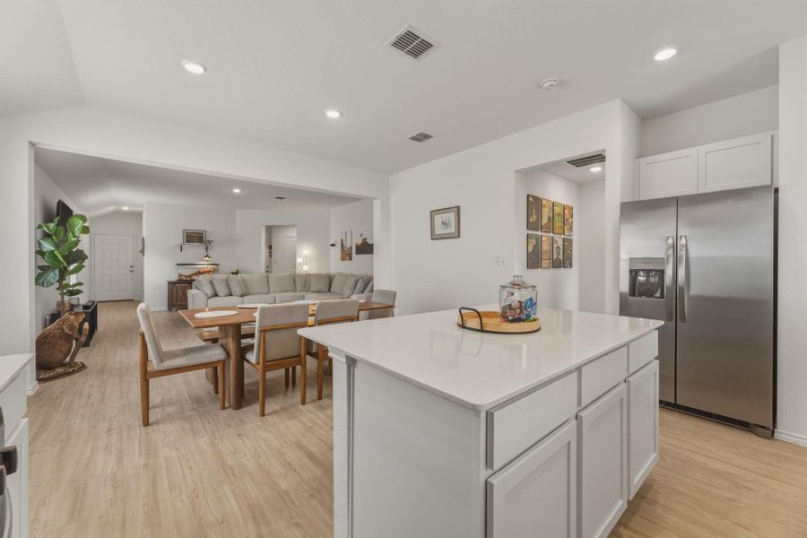 The kitchen features a large island with a light-colored countertop and white cabinetry, recessed lighting, and a stainless steel refrigerator