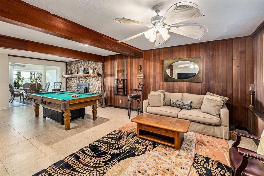 Spacious recreation area featuring wood paneling, a stone fireplace, exposed wood beams, tile flooring, and a ceiling fan with integrated lighting