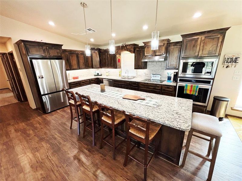 Kitchen with dark brown cabinets, decorative light fixtures, stainless steel appliances, light stone countertops, and a breakfast bar area Kitchen with dark brown cabinets, decorative light fixtures, stainless steel appliances, light stone countertops, and a breakfast bar area