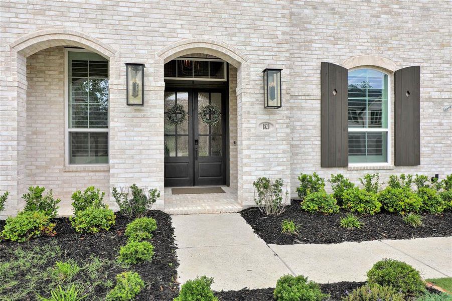 Exterior featuring light-toned brick, arched entryways, dark double doors, rectangular lantern-style sconces, and dark wood shutters