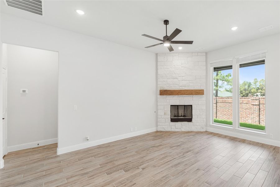 Open living room with stone fireplace, wood mantel, ceiling fan, and light wood-look floors