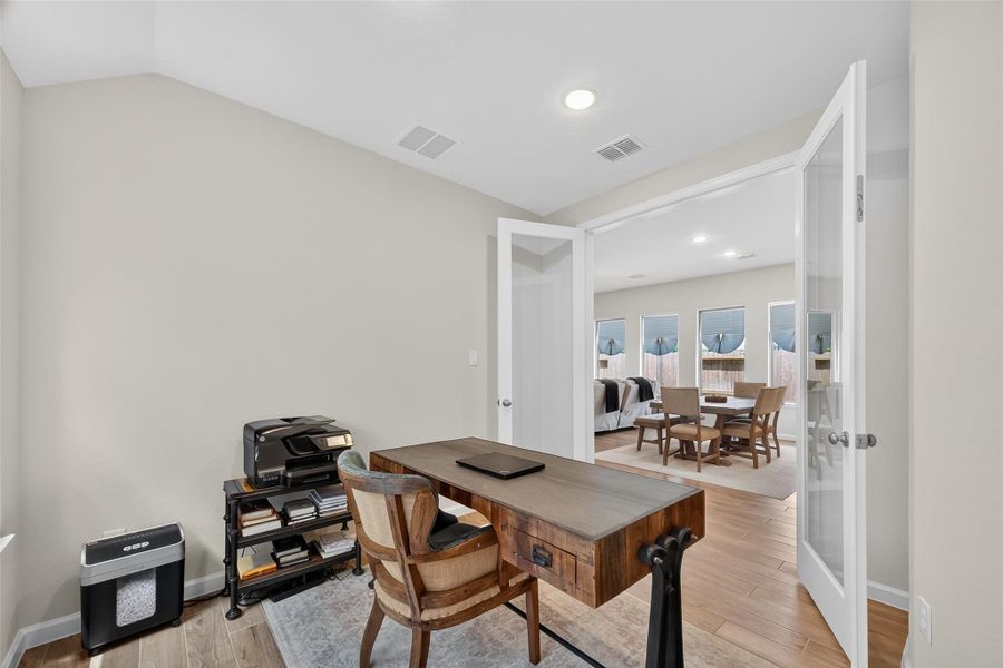 This photo shows a bright home office with wood flooring, a desk, and a chair. It features double glass doors leading to a spacious room with multiple windows, providing ample natural light. This photo shows a bright home office with wood flooring, a desk, and a chair. It features double glass doors leading to a spacious room with multiple windows, providing ample natural light.