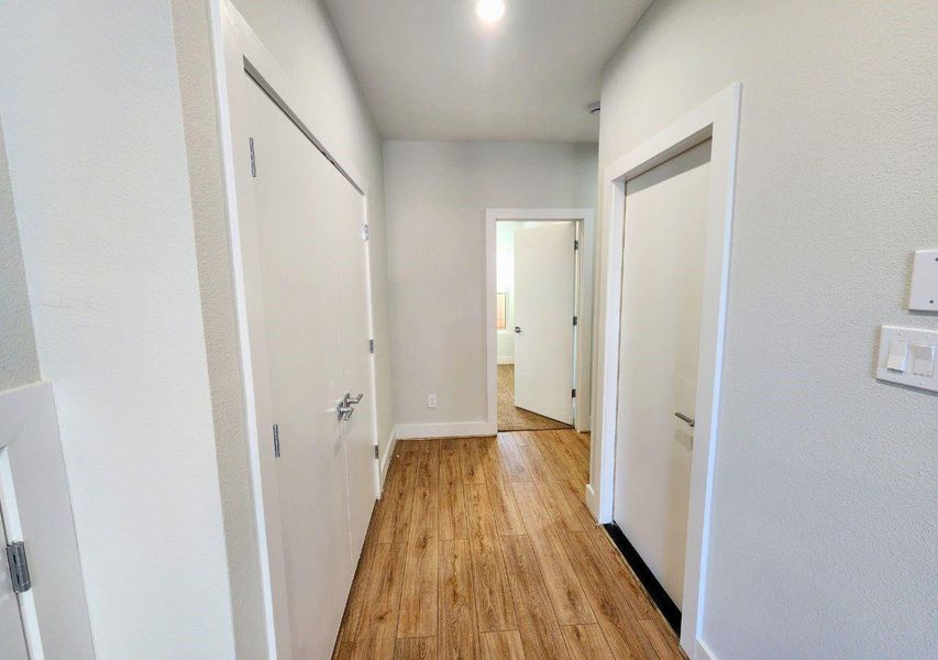 This photo shows a bright hallway with wood-style flooring and neutral painted walls. There are two closed doors on the left and one on the right, leading to another room at the end. This photo shows a bright hallway with wood-style flooring and neutral painted walls. There are two closed doors on the left and one on the right, leading to another room at the end.