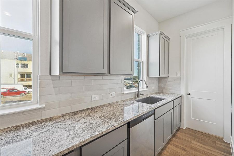 Kitchen with gray cabinetry, light wood-style flooring, stainless steel dishwasher, backsplash, and light stone countertops