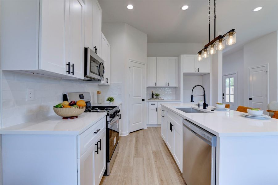 Full kitchen showing floor-to-ceiling white shaker cabinets with matte black hardware, quartz countertops, subway tile backsplash, stainless gas range, over-the-range microwave, stainless dishwasher, farmhouse sink on island, and Edison rail pendant. Pantry door and rear exit visible at end of kitchen.