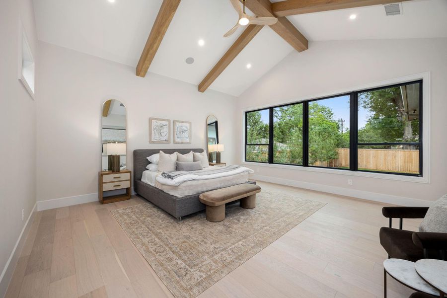 A serene and spacious primary bedroom designed for comfort and relaxation, featuring wide-plank flooring and a soft, neutral palette. Large picture windows flood the room with natural light while framing views of the outdoors. The highlight of this space is the vaulted ceiling with exposed wood beams, adding architectural character and warmth while creating an open, airy retreat that feels both luxurious and inviting.