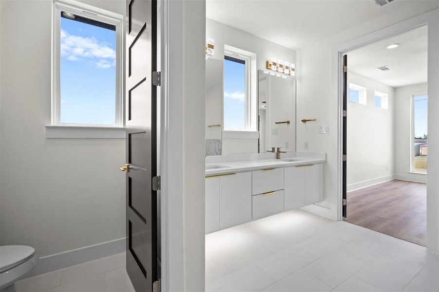 Bright bathroom featuring a dual vanity with white cabinetry, gold hardware, and a modern light fixture Bright bathroom featuring a dual vanity with white cabinetry, gold hardware, and a modern light fixture