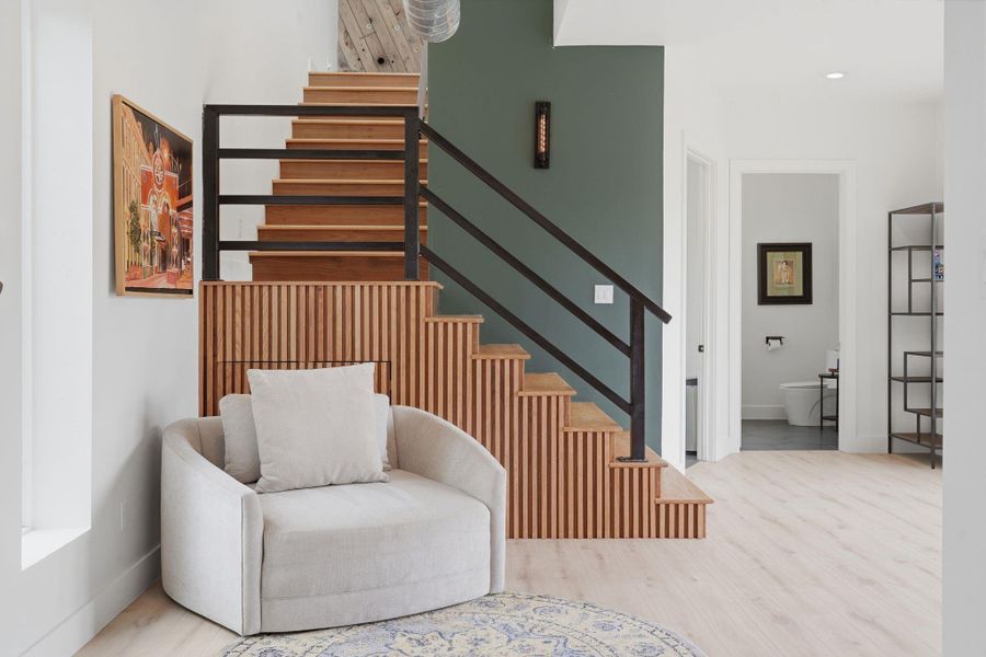 This photo showcases a modern interior with a stylish wooden staircase (with built in storage) featuring black railings. The neutral-toned room includes a cozy armchair and decorative elements like a wall painting and a minimalist shelving unit. The space feels open and inviting, with light wood flooring and a small glimpse into an adjacent room.