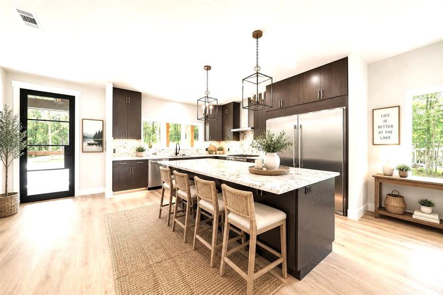 Virtually staged-Spacious kitchen featuring an oversized island with countertop seating, dark wood-finish cabinetry, stainless steel appliances, light wood-finish flooring, and a glass-paned door Virtually staged-Spacious kitchen featuring an oversized island with countertop seating, dark wood-finish cabinetry, stainless steel appliances, light wood-finish flooring, and a glass-paned door