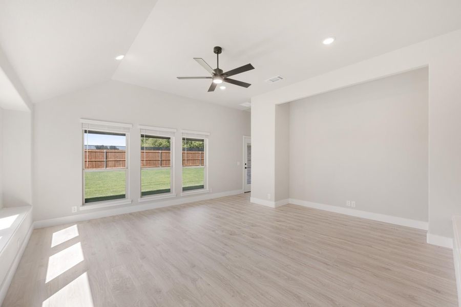 Empty living room with vaulted ceiling, ceiling fan, light wood floors, and three windows overlooking backyard