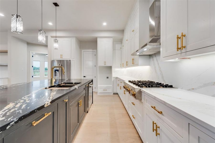 Kitchen featuring wall chimney exhaust hood, a sink, stainless steel gas stovetop, white cabinetry, and light stone counters Kitchen featuring wall chimney exhaust hood, a sink, stainless steel gas stovetop, white cabinetry, and light stone counters
