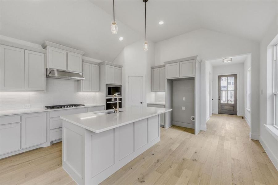 Kitchen featuring light wood-style floors, pendant lighting, a center island with sink, lofted ceiling, and backsplash Kitchen featuring light wood-style floors, pendant lighting, a center island with sink, lofted ceiling, and backsplash