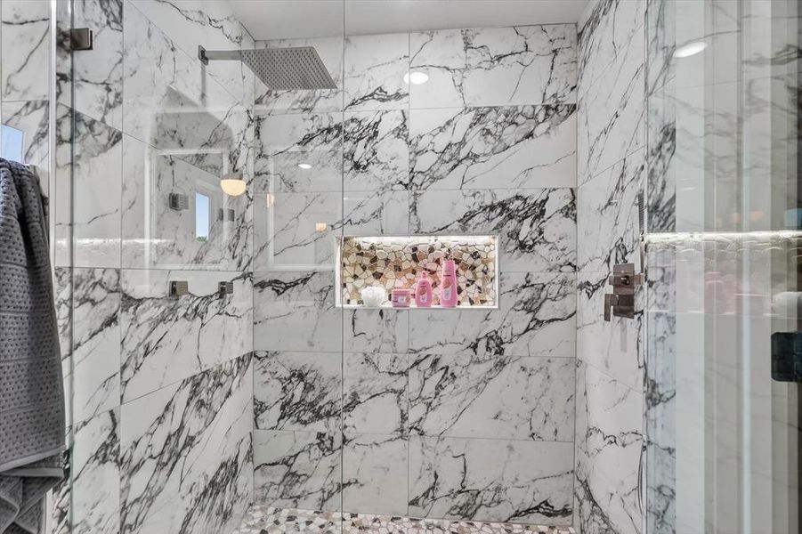 Spacious shower featuring white marble-look wall tiling, a pebble-style floor, a built-in illuminated niche, and a rain showerhead
