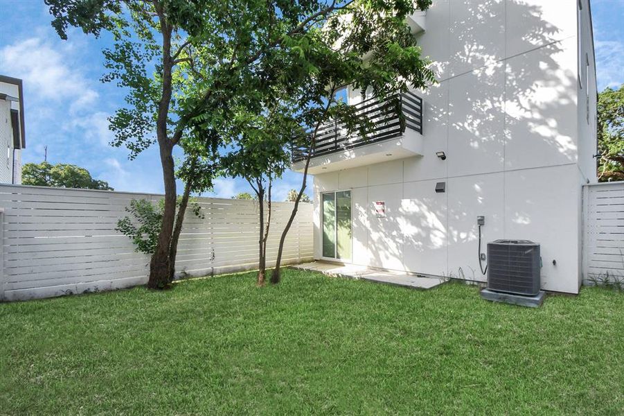 This is a modern two-story home featuring a clean white exterior with a balcony on the upper level. The property includes a private fenced yard with a well-maintained lawn and mature trees, providing a blend of open space and natural shade. The home comes with an outdoor air conditioning unit and offers a combination of privacy and contemporary living.