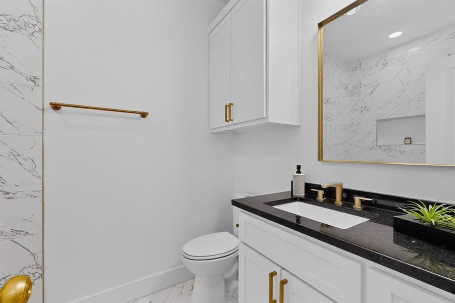 White vanity with a black countertop, gold-tone hardware, and a framed mirror