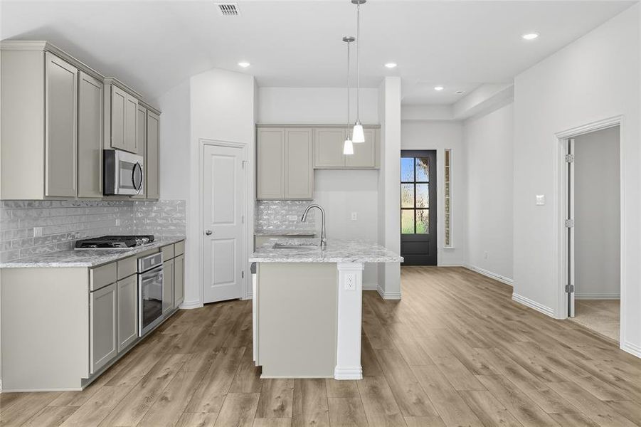 Kitchen featuring light stone counters, tasteful backsplash, a kitchen island with sink, pendant lighting, and light wood-type flooring Kitchen featuring light stone counters, tasteful backsplash, a kitchen island with sink, pendant lighting, and light wood-type flooring