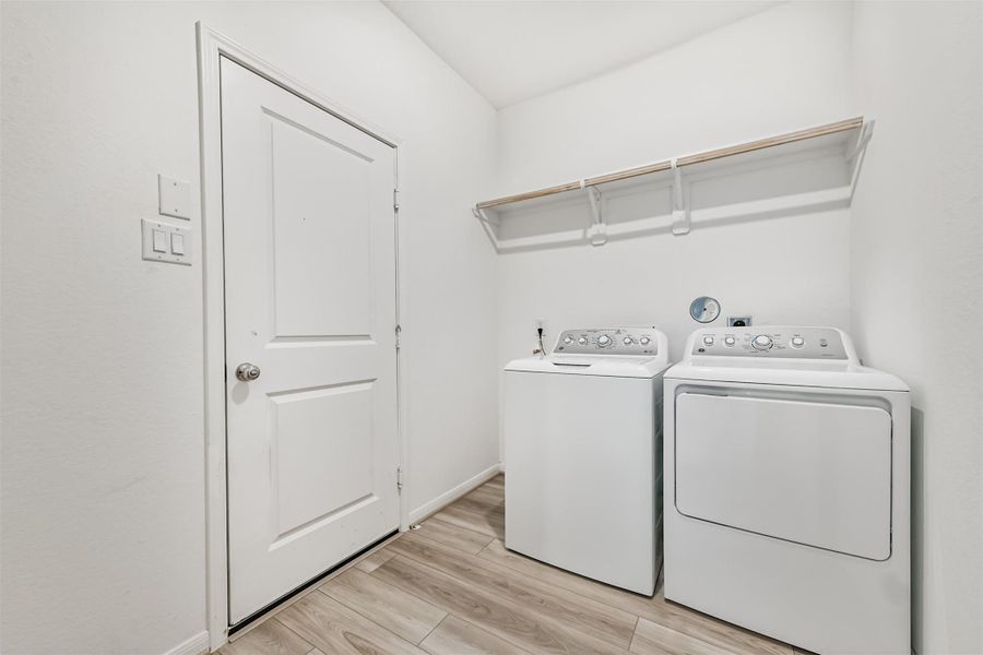The bright laundry room with washer and dryer included and a wooden shelf above for storage. The door leads out to the two car garage where the tankless water heater is located.