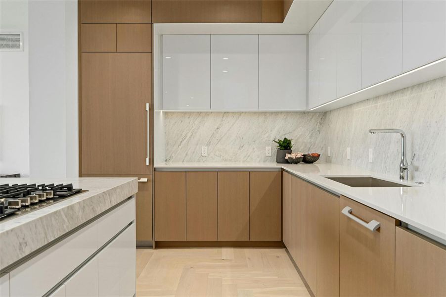 The kitchen features a sleek, minimalist design with a large island at its center, topped with marble stone countertop. It includes a built-in gas cooktop. Custom cabinetry seamlessly conceals the integrated refrigerator, maintaining clean lines and a refined aesthetic.