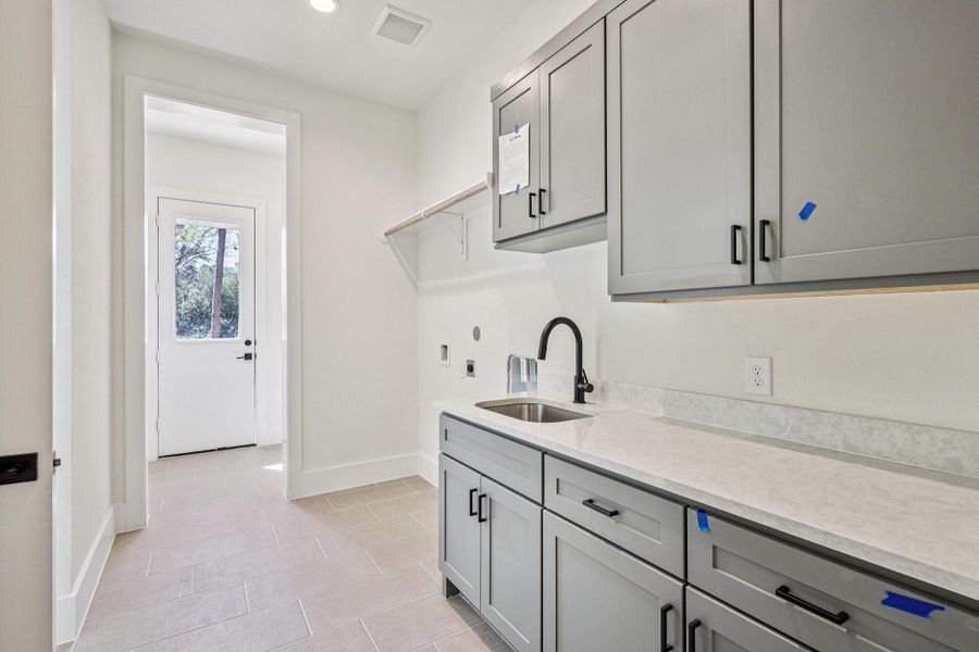 This photo showcases a modern laundry room with sleek gray cabinets, a countertop, and a sink. The space is well-lit and features tile flooring, with a door leading outside, making it both functional and stylish.