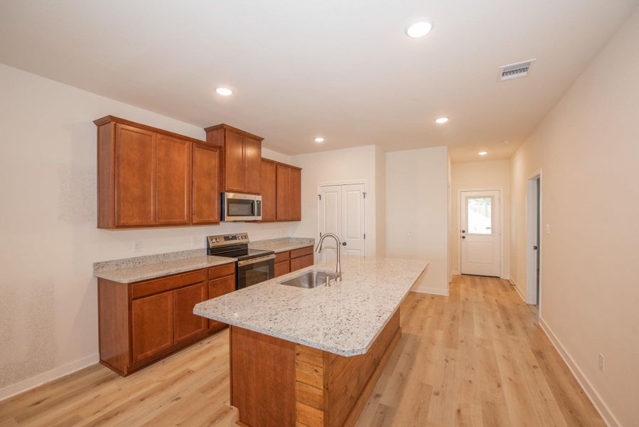 Kitchen featuring brown cabinets, appliances with stainless steel finishes, light wood-type flooring, light stone countertops, and a kitchen island with sink