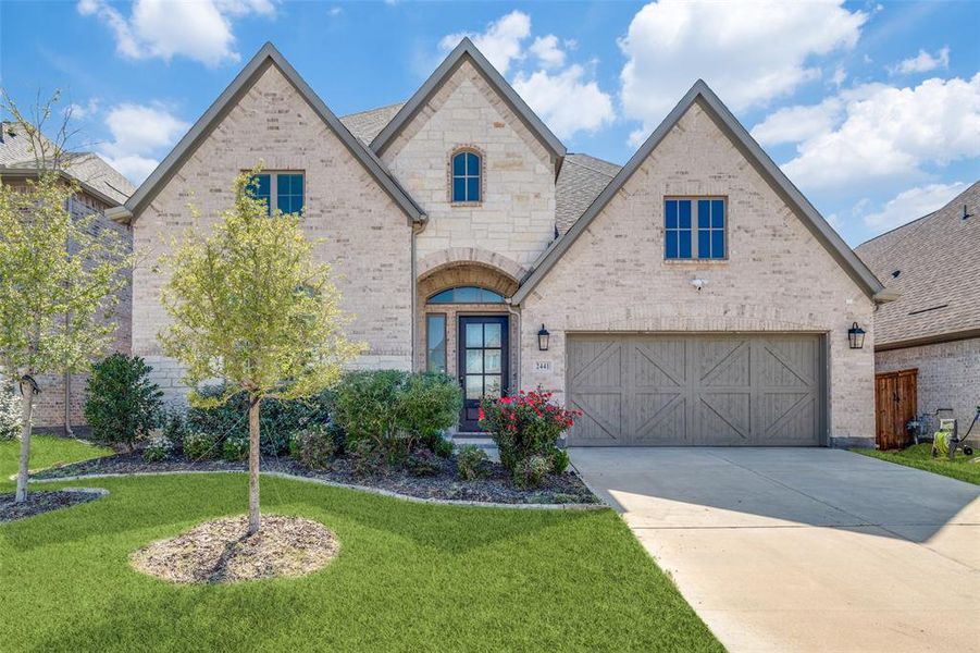French country inspired facade with brick siding, driveway, a garage, a front lawn, and stone siding