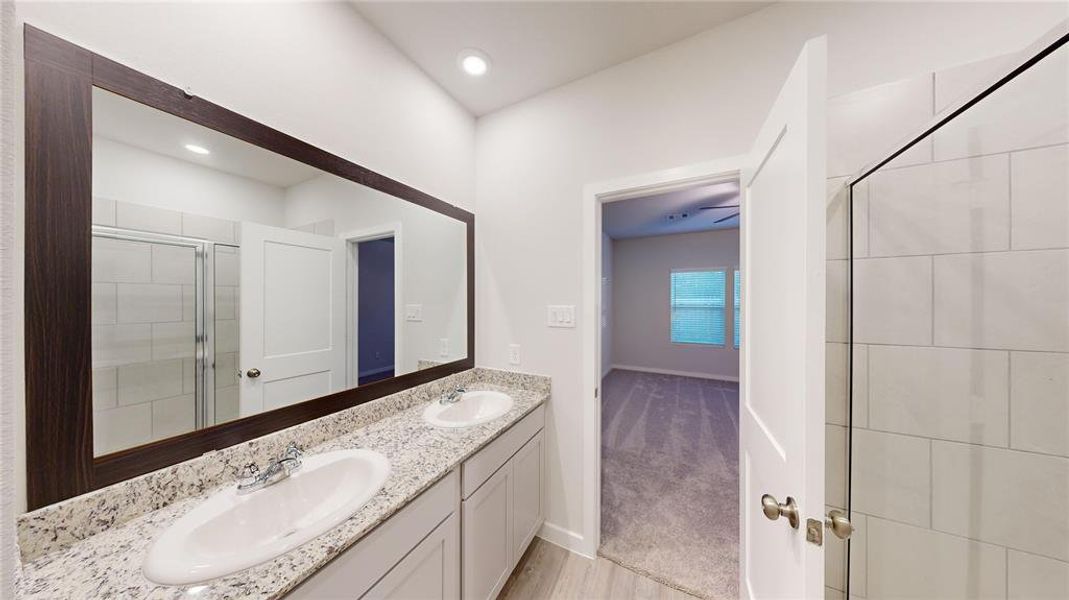 This photo features a modern bathroom with a double sink vanity, granite countertops, and a large mirror. It includes a tiled shower with a glass door and opens to the primary bedroom.