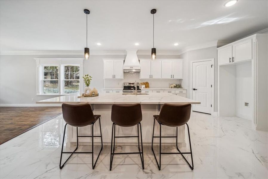 Kitchen featuring custom exhaust hood, an island with sink, hanging light fixtures, white cabinets, and light stone counters