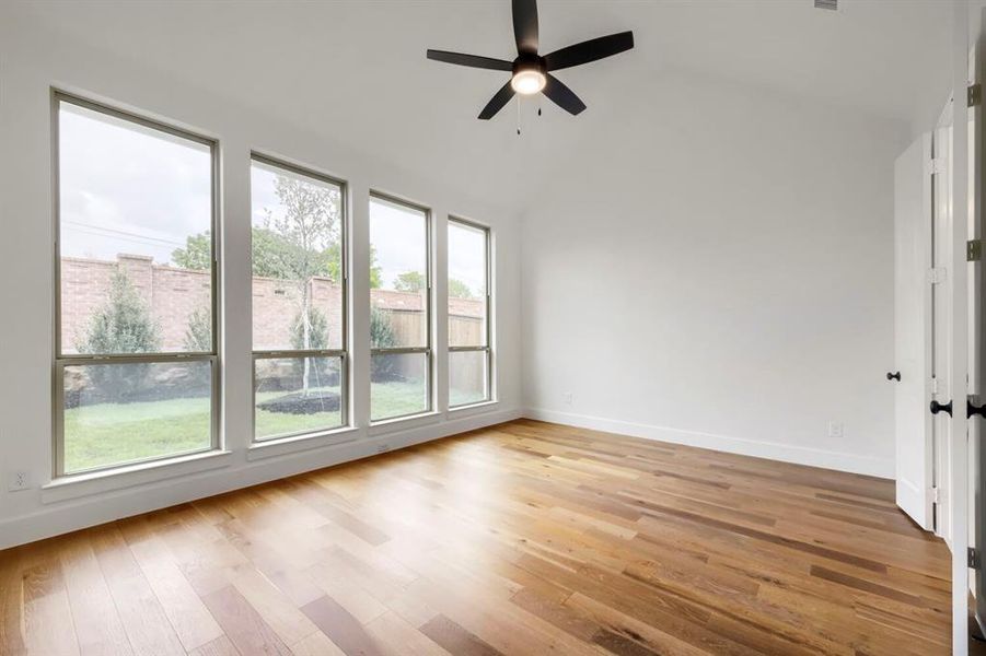 This room features hardwood flooring, a vaulted ceiling with a ceiling fan, and large windows that provide views of the yard
