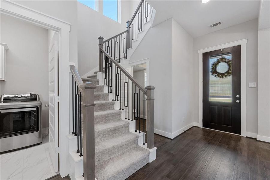 This is a modern entryway featuring laminate hardwood floors, white walls, and an elegant staircase with a wooden handrail and black iron spindles. A front door with glass panels allows natural light to enter, and the space feels open and welcoming.