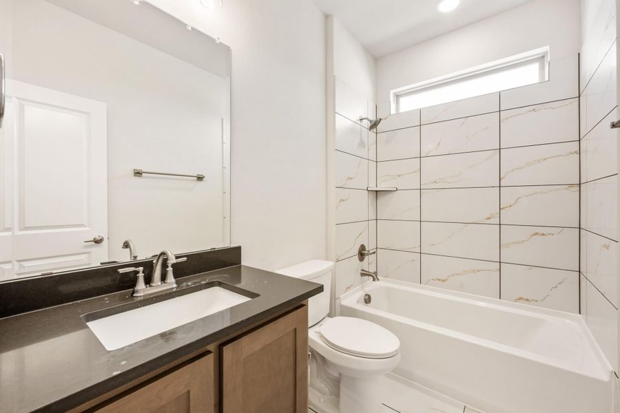 Bathroom with dark quartz vanity, wood cabinets, and tub-shower combo with marble-look tile surround Bathroom with dark quartz vanity, wood cabinets, and tub-shower combo with marble-look tile surround