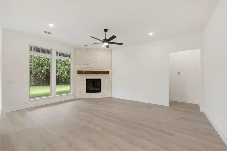 Empty living room with stone fireplace, wood mantel, ceiling fan, and light hardwood floors