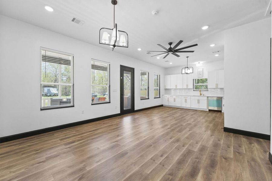 This spacious, open-concept room features modern lighting and a sleek ceiling fan. The kitchen area includes white cabinetry, a stylish backsplash, and stainless steel appliances. Large windows provide ample natural light, enhancing the bright and airy feel.