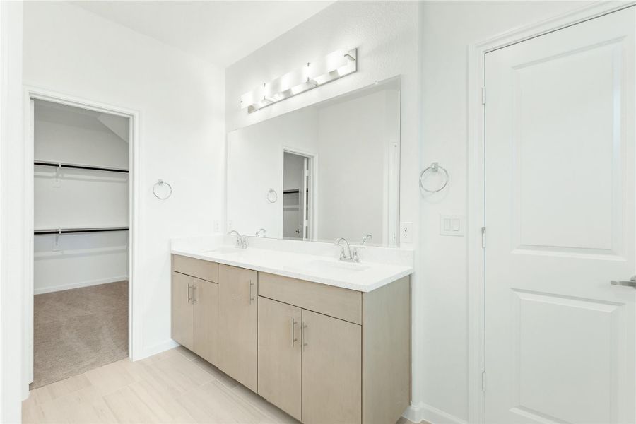 Bathroom with double sink vanity, tan cabinets, large mirror, and walk-in closet visible through doorway