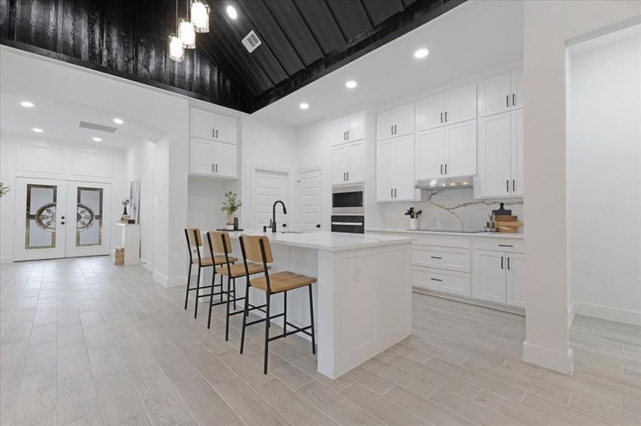 Kitchen with tasteful backsplash, a kitchen island with sink, light countertops, under cabinet range hood, and white cabinetry